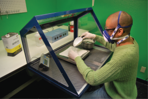 Above, a technician weighs silica on the perforated bench top of a Model 435 Portable Downdraft Bench - Sit, which pulls renegade particles down into filters that trap them below the work surface, away from his mouth and nose.